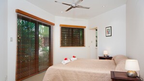 Un dormitorio con una cama, dos ventanas con persianas, un ventilador de techo, una mesita de noche con lámpara y un cuadro en la pared.