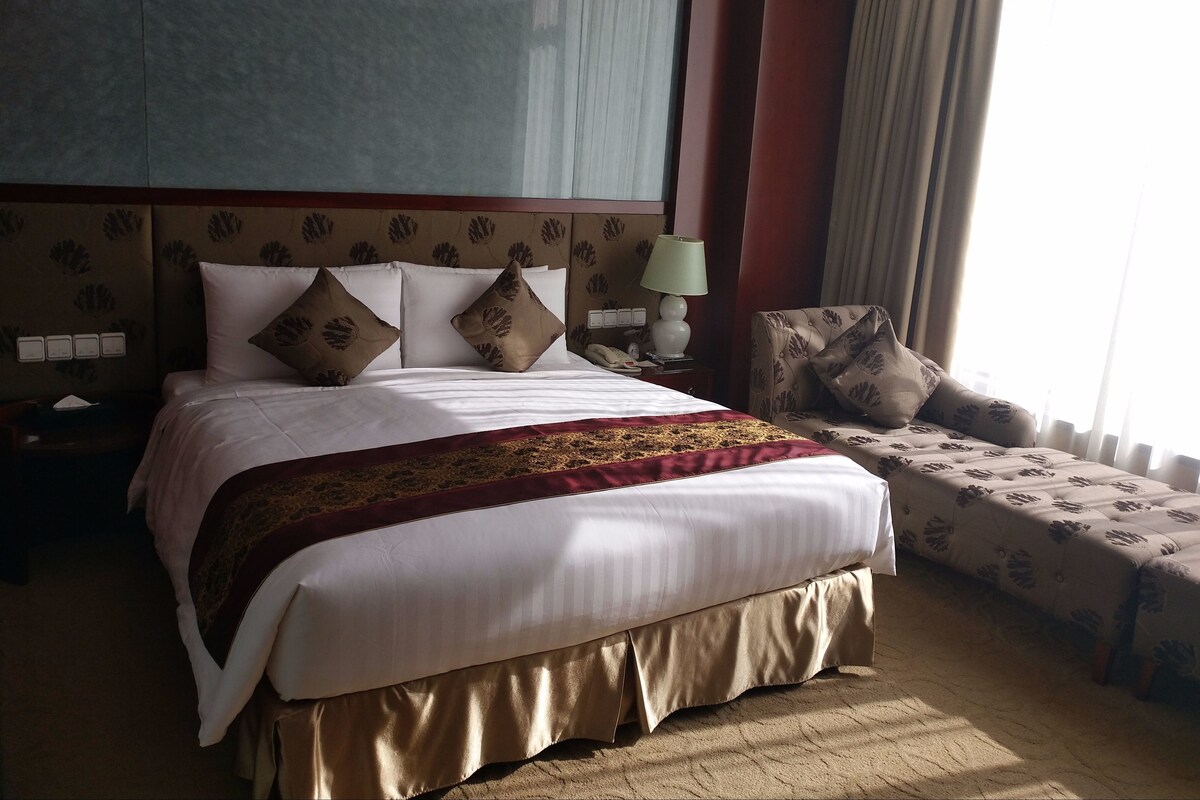 Ein Hotelzimmer mit einem großen Bett, einem Nachttisch mit Lampe, einem Sessel und einem großen Fenster mit Vorhängen.