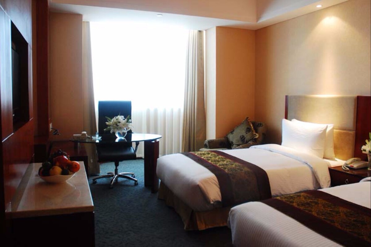 Ein Hotelzimmer mit zwei Betten, einem Schreibtisch mit Stuhl, einem kleinen Tisch mit Blumenvase und Blick ins Freie durch ein Fenster.