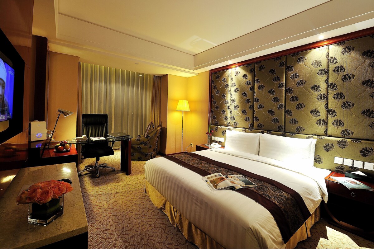 Ein Hotelzimmer mit einem Bett, einem Schreibtisch mit Laptop, einem Sessel, einem Fernseher und einer Lampe.
