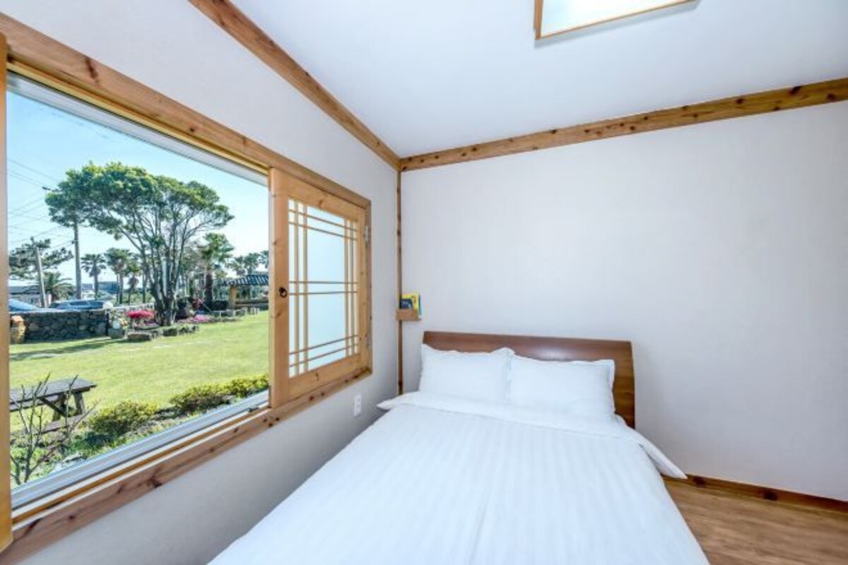 A bedroom with a wooden headboard, a large window with a view of a garden, and a neatly made bed.