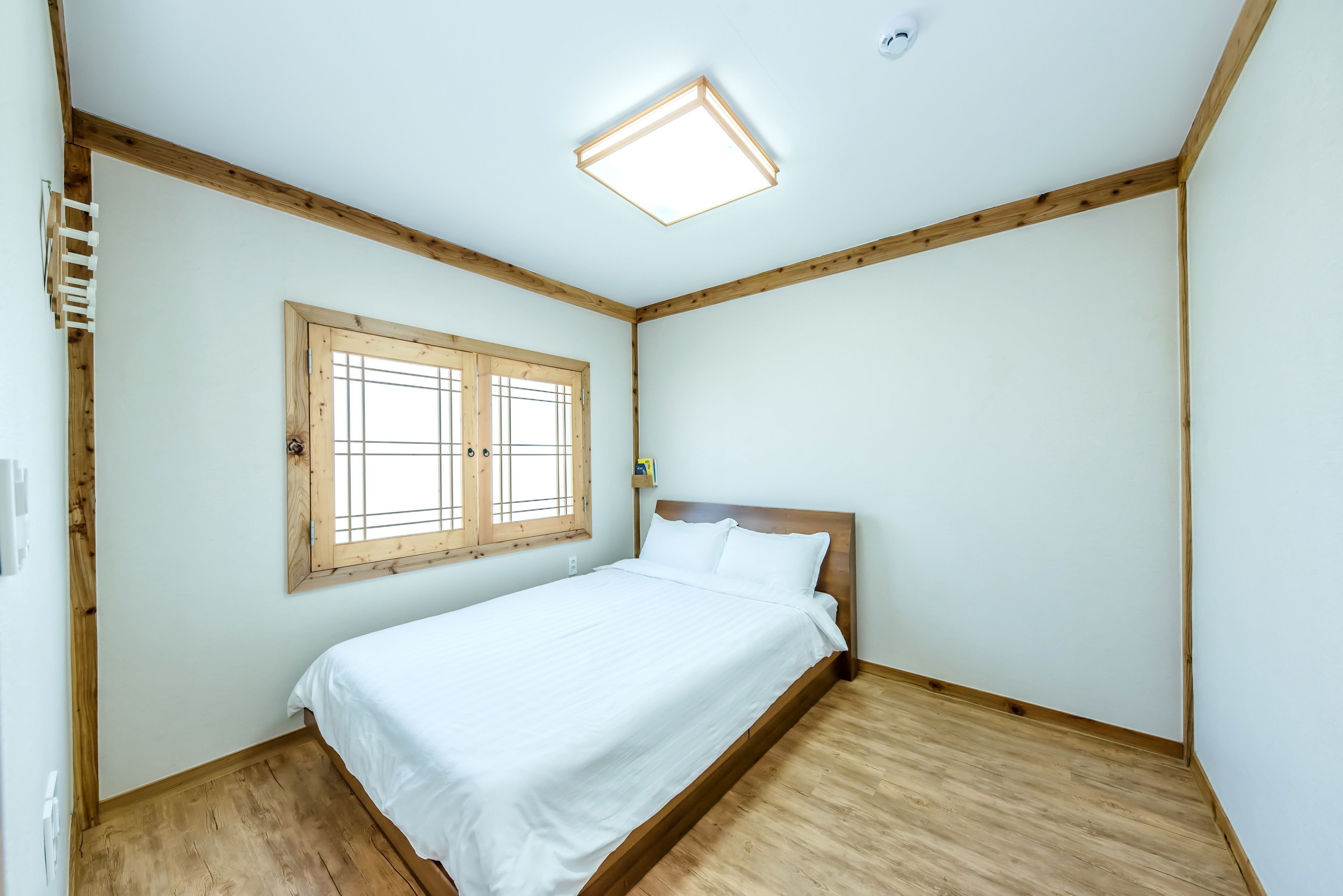 A bedroom with a wooden floor, a bed with white bedding, a window with wooden frames, and a ceiling light fixture.