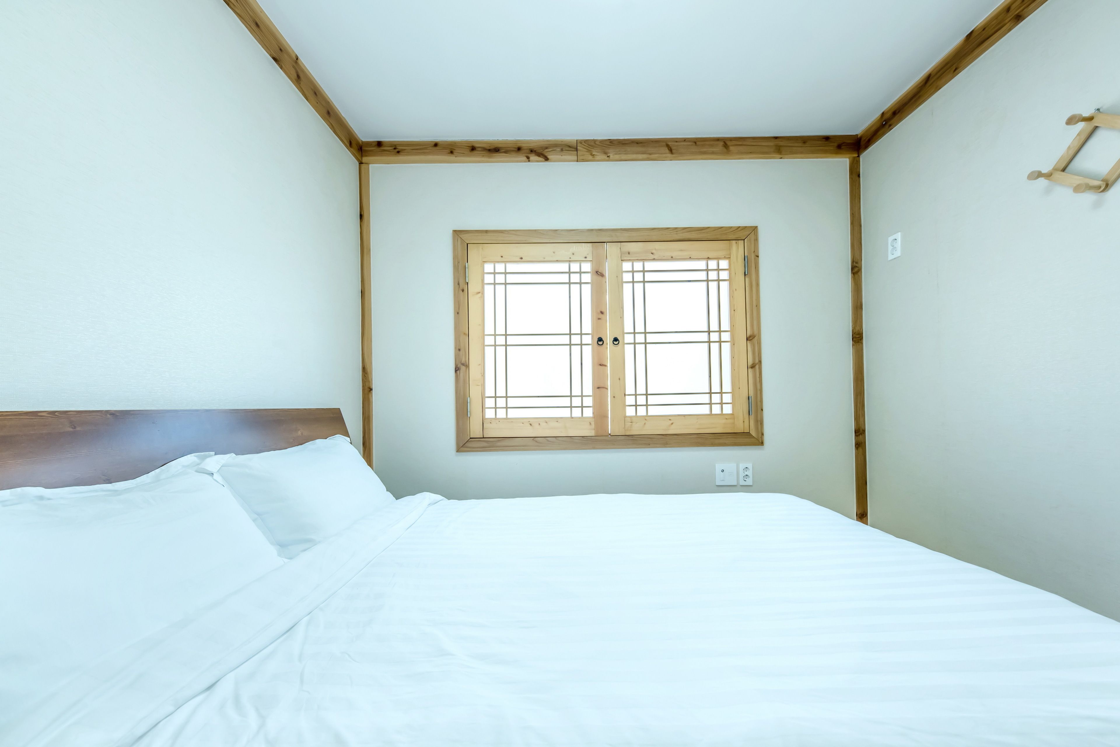Een slaapkamer met een houten hoofdbord, een groot raam met houten kozijnen en een wit bed met kussens.