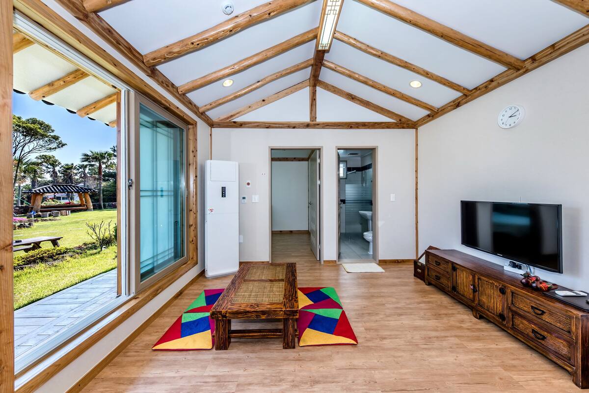 A spacious living room with a large window, wooden ceiling beams, a television, and a dining area.
