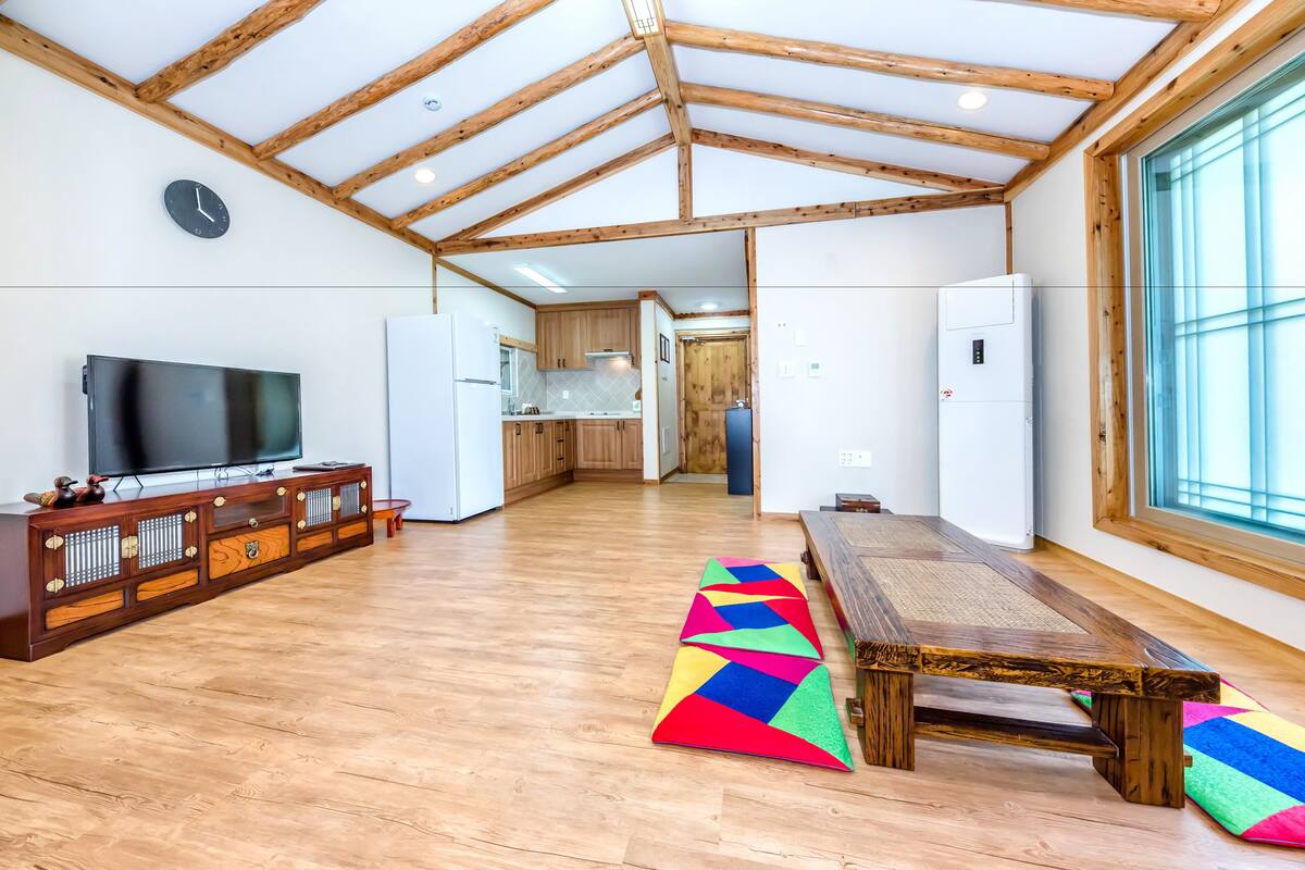 A spacious living area with a wooden ceiling, a large flat-screen TV, a dining table, and colorful mats on the floor.