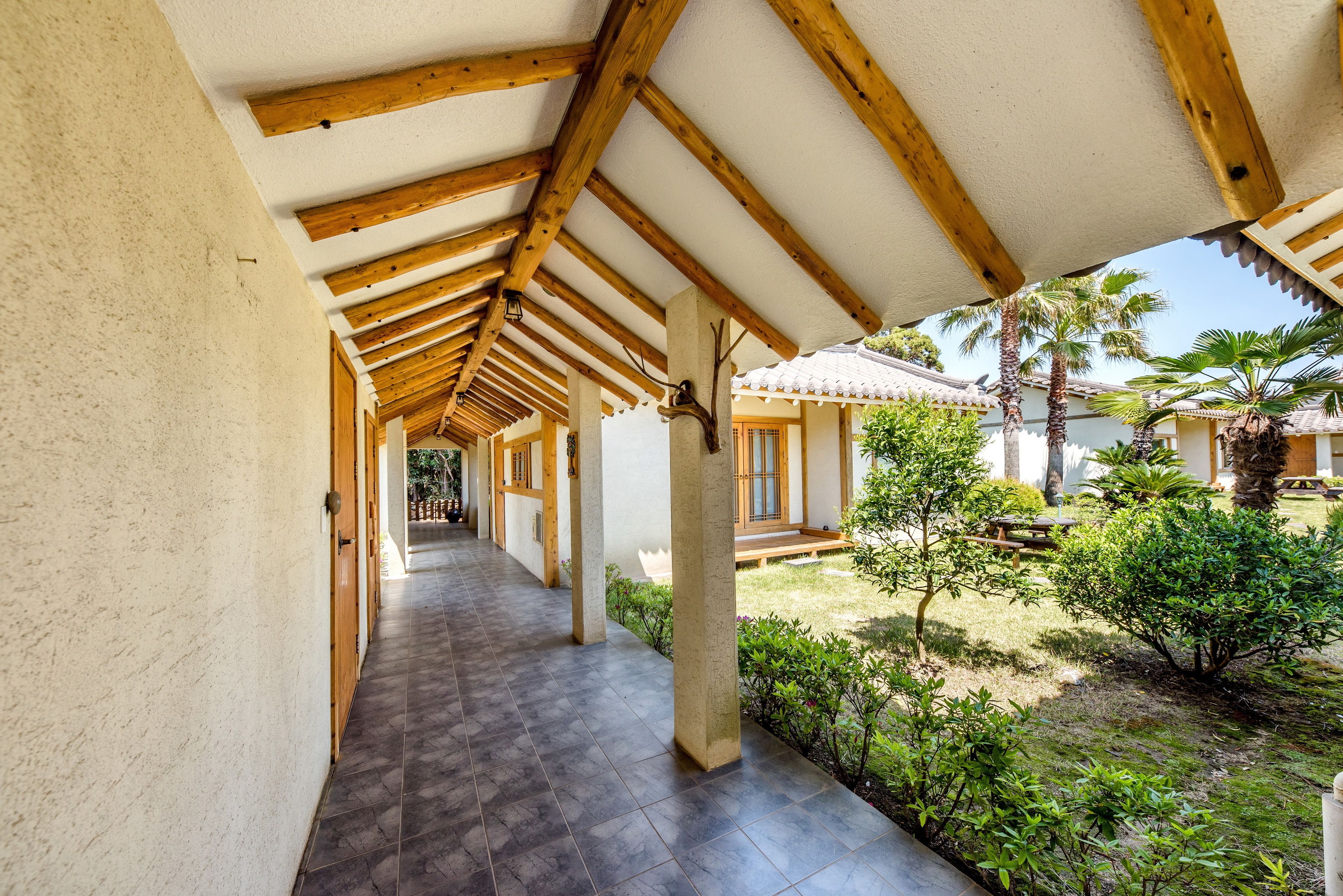 A covered corridor with wooden beams and tiled flooring, leading to a courtyard with palm trees and a traditional building with tiled roof.