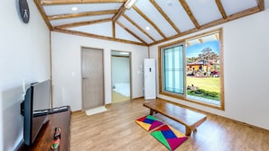 A spacious room with wooden beams, a large window, a television, and a colorful rug. - Stone Statue Village Hanok (Seogwipo)