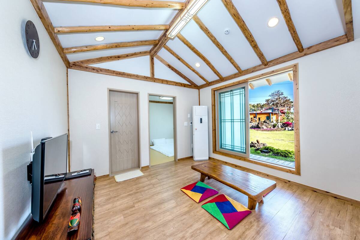 A spacious room with wooden beams, a large window, a television, and a colorful rug.