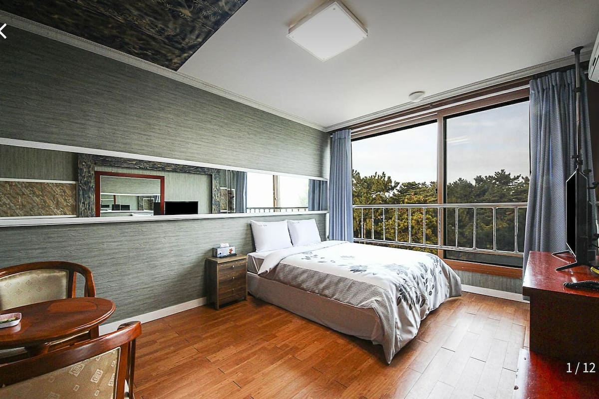 Ein Schlafzimmer mit einem großen Bett, einem Holztisch, einem Stuhl, einem Nachttisch, einem Spiegel und einem Fenster mit Blick auf Bäume.