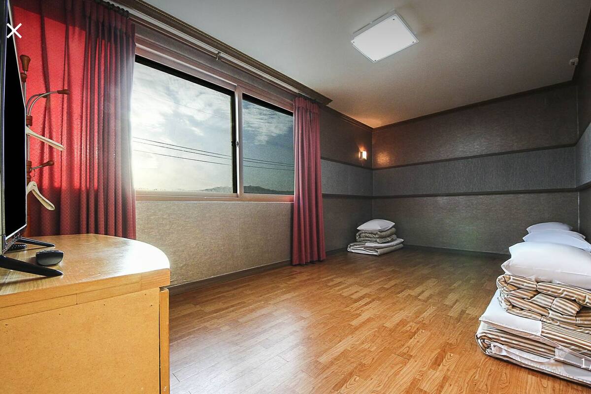 Ein Hotelzimmer mit Holzboden, einem Schreibtisch mit Fernseher, einem Fenster mit roten Vorhängen und einem Bett mit Kissen und gefalteter Kleidung.