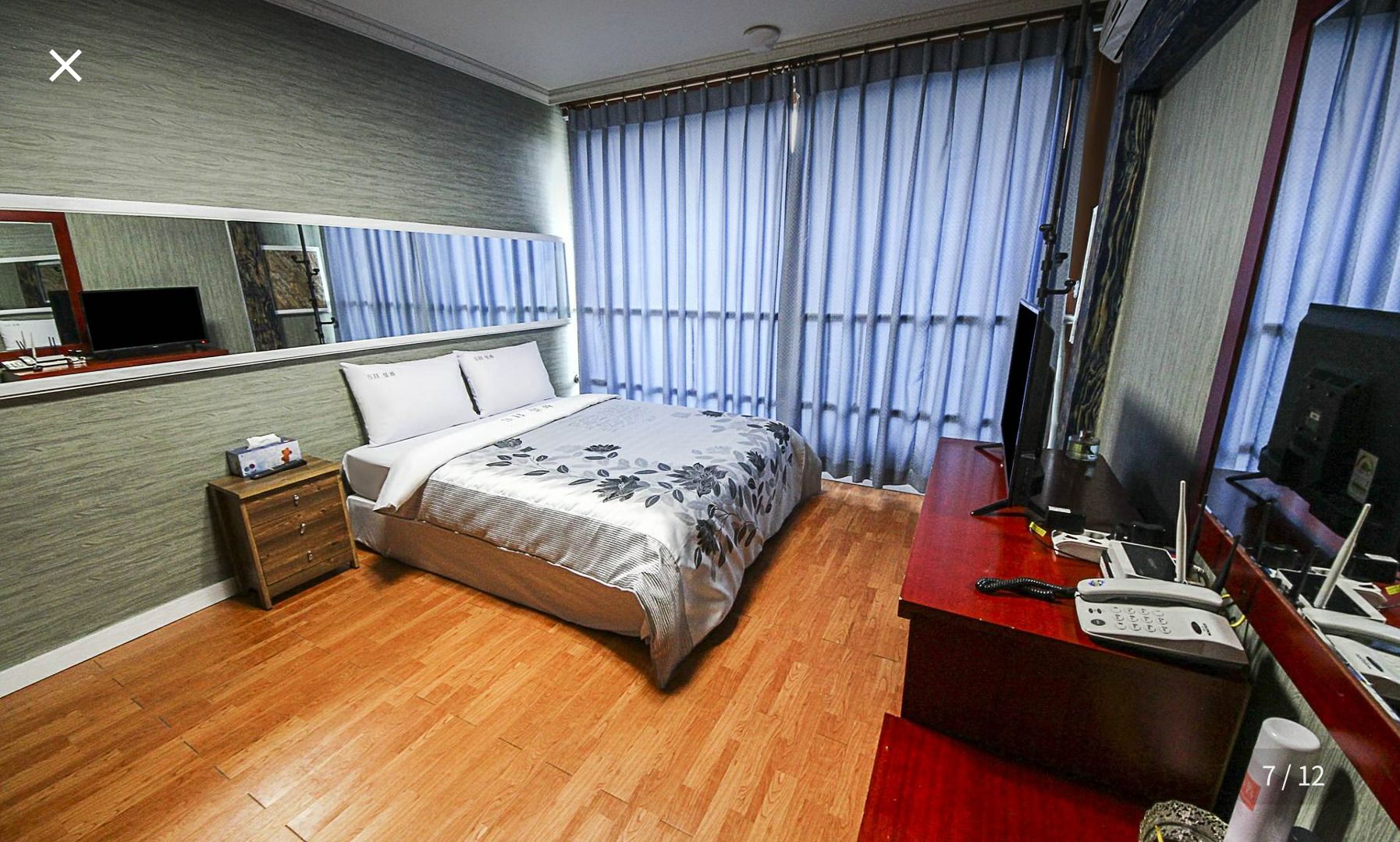 Quarto de hotel com piso de madeira, cama com colcha estampada, escrivaninha com telefone e um espelho grande.