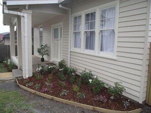 A house with a covered porch, a garden bed with various plants, and a window with white curtains. - Avenue House (Moonah)