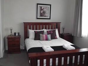 A neatly made bed with white linens and multiple pillows, a wooden headboard, a bedside table with a lamp, and a framed picture on the wall. - Avenue House (Moonah)
