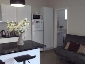 A modern kitchen with white cabinets, a black countertop, a microwave, and a refrigerator. There is a vase with white flowers on the countertop. - Avenue House (Moonah)