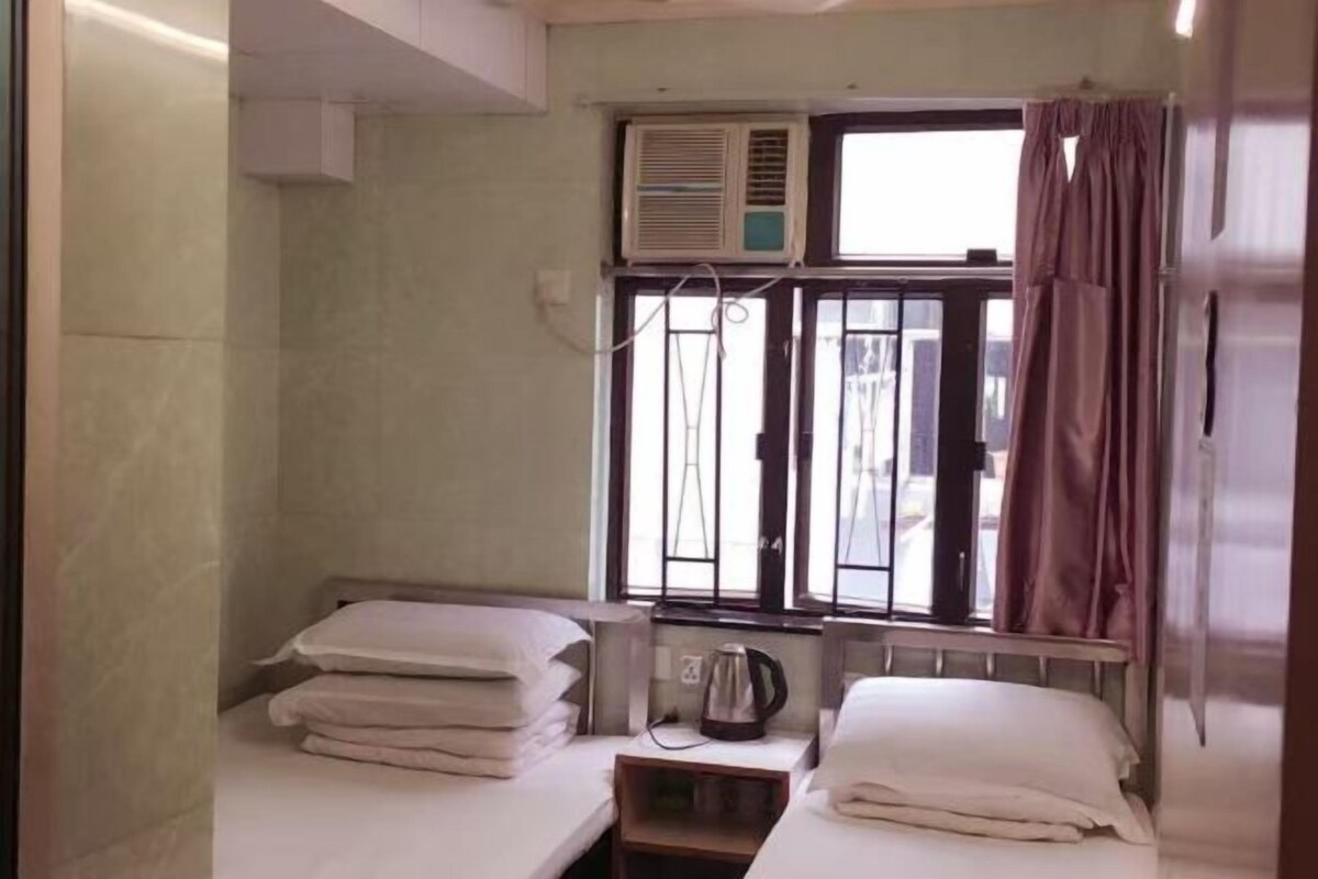 A small, single-room hotel room with two beds, a window with bars, an air conditioning unit, and a kettle on the nightstand.
