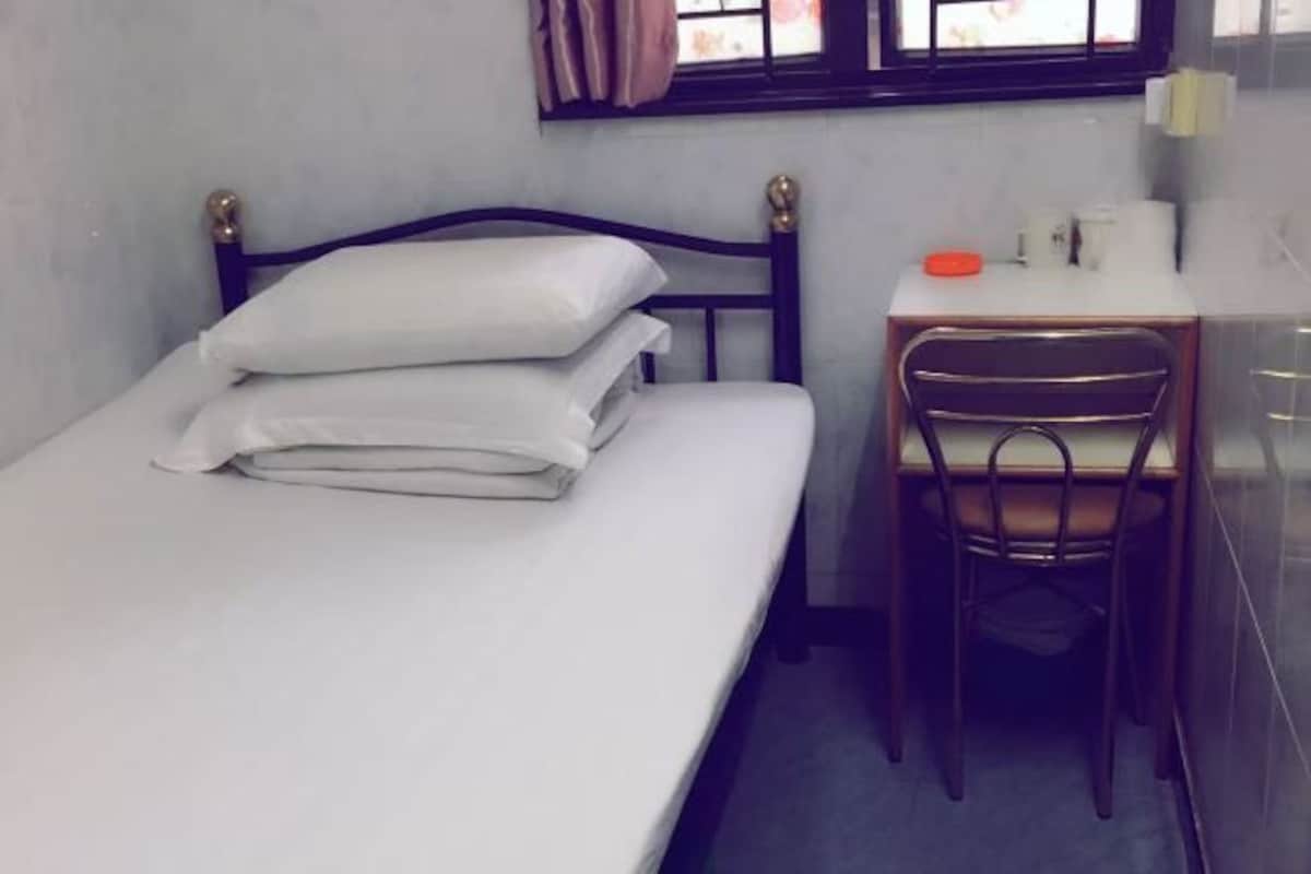 Quarto pequeno com uma cama de solteiro, cabeceira de madeira, uma mesa com cadeira e uma janela com cortinas.