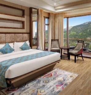 A hotel room with a large bed, a balcony with a view of the mountains, a small table, and a chair. - Alokik Resort (Manali)