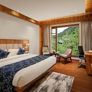A hotel room with a large bed, two chairs, a desk, and a view of a forested landscape. - Alokik Resort (Manali)