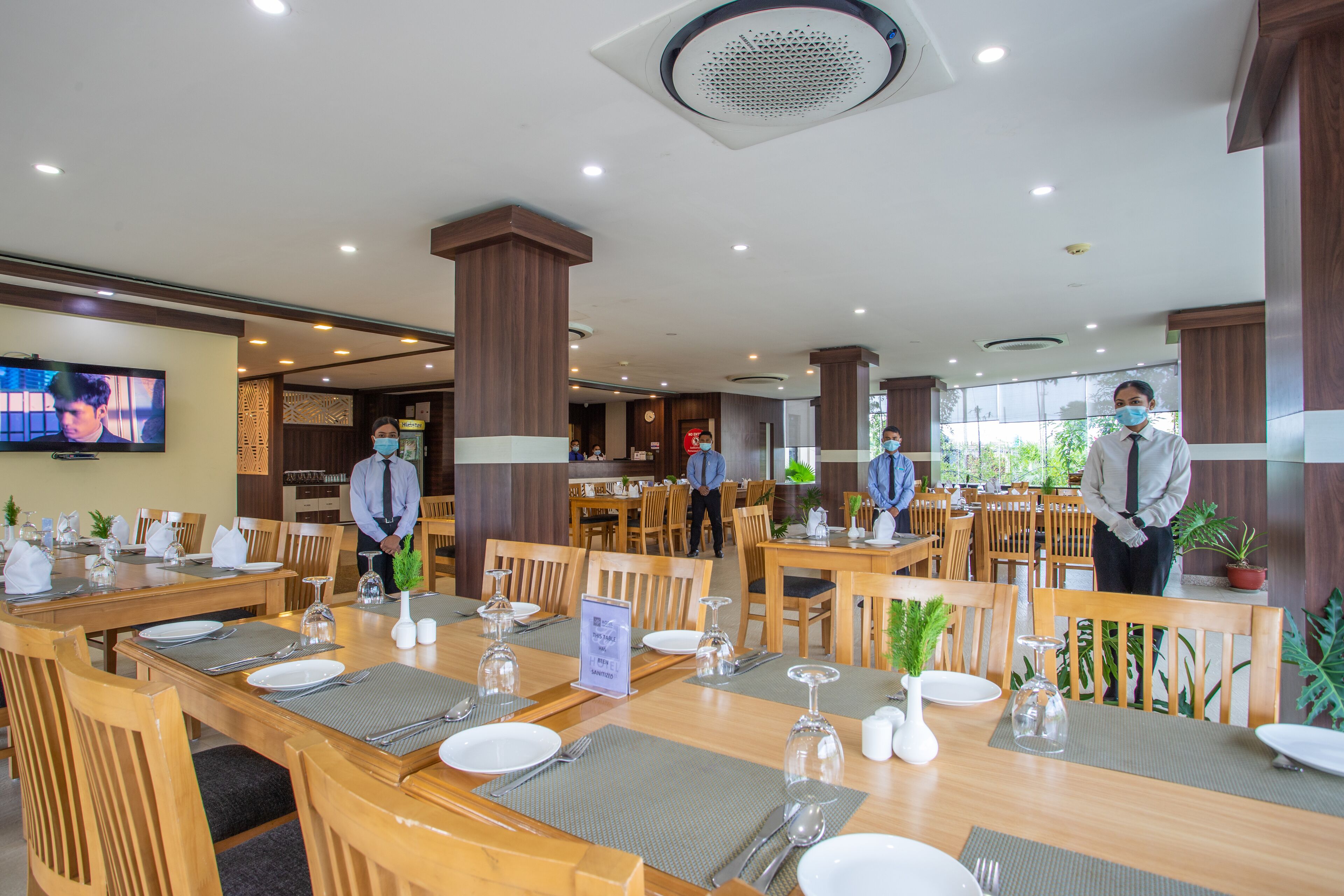 A hotel dining area with multiple tables set for guests, wooden chairs, and a television screen on the wall.