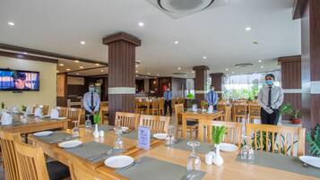 A hotel dining area with multiple tables set for guests, wooden chairs, and a television screen on the wall.