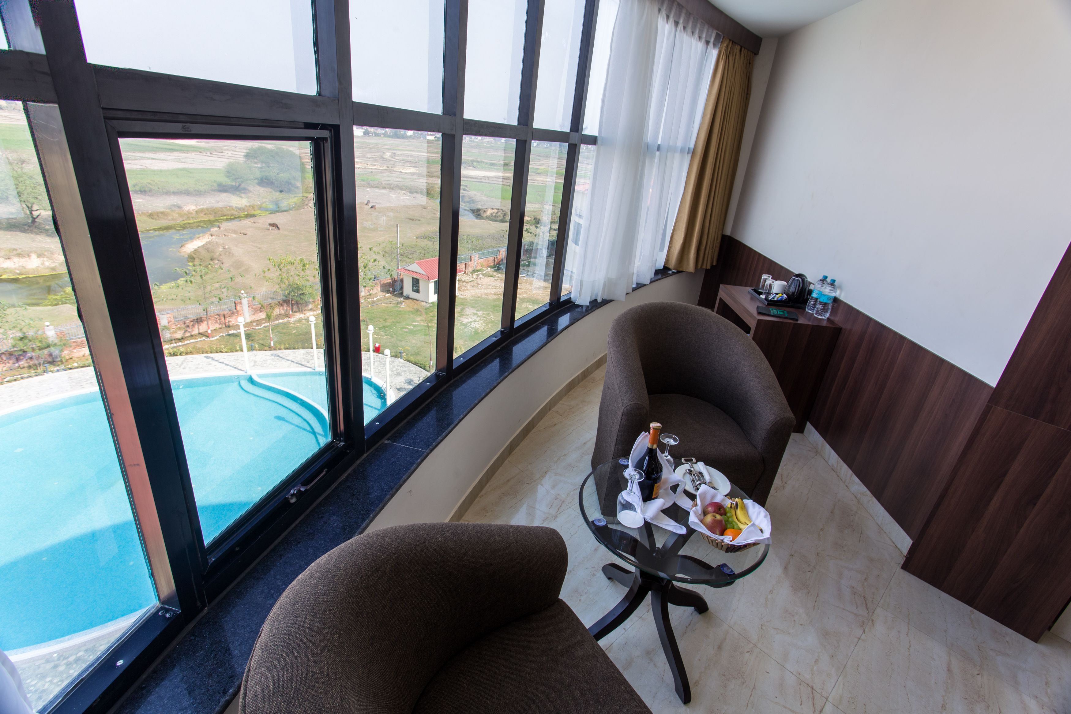 Un balcón con vista a una piscina y el paisaje, un sillón marrón, una mesita con una botella y una botella de agua en una mesita de noche.