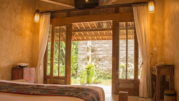 Un dormitorio con cama, muebles de madera, una mesita y vistas a la vegetación a través de una puerta abierta.