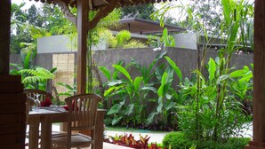 Un patio al aire libre con una mesa y sillas de madera, rodeado de abundante vegetación y un muro de piedra.