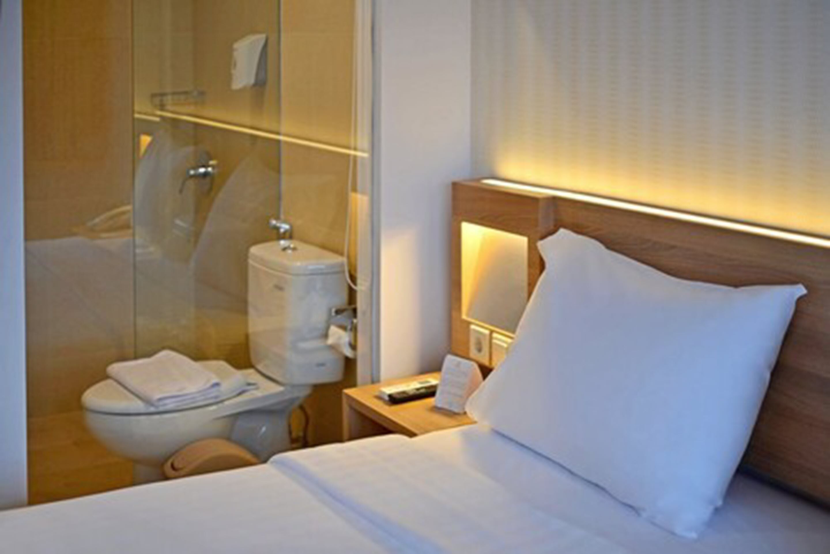 Habitación de hotel con cama, mesita de noche con lámpara, cabina de ducha de vidrio y retrete.