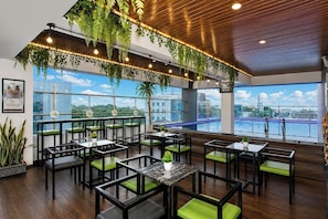 A restaurant with outdoor seating, green cushioned chairs, hanging plants, and a view of a water body. - Whiz Prime Hotel Basuki Rahmat Malang (Malang)