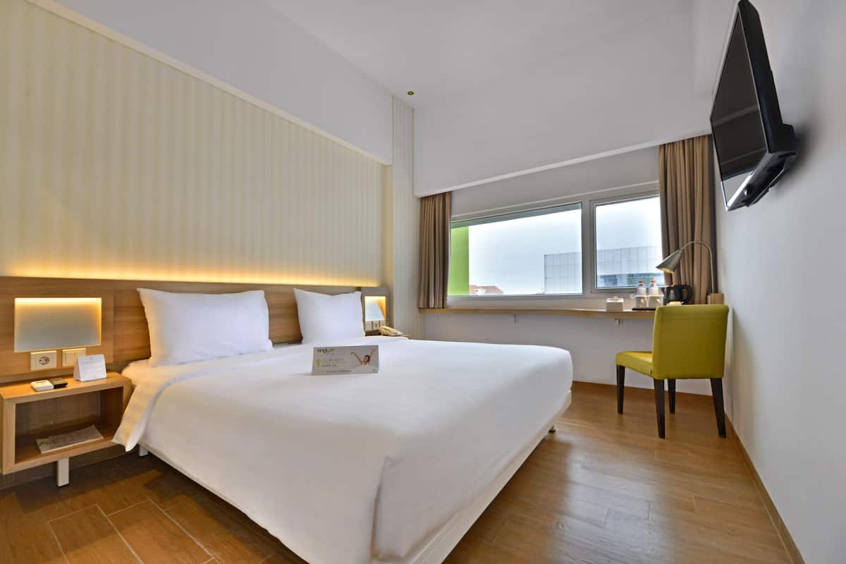 A hotel room with a large bed, a bedside table with a lamp, a TV mounted on the wall, and a window with curtains.