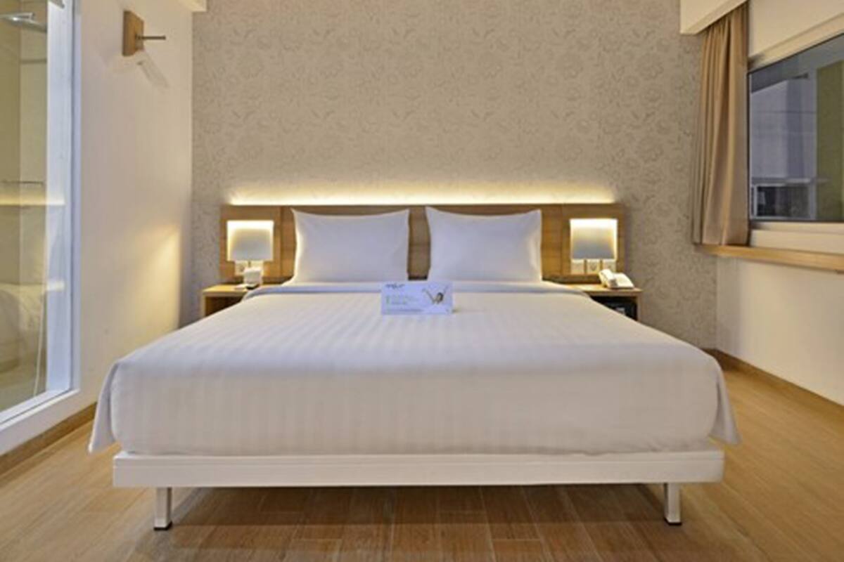 A hotel room with a large bed, two bedside tables with lamps, a window with curtains, and a wall with a subtle pattern.