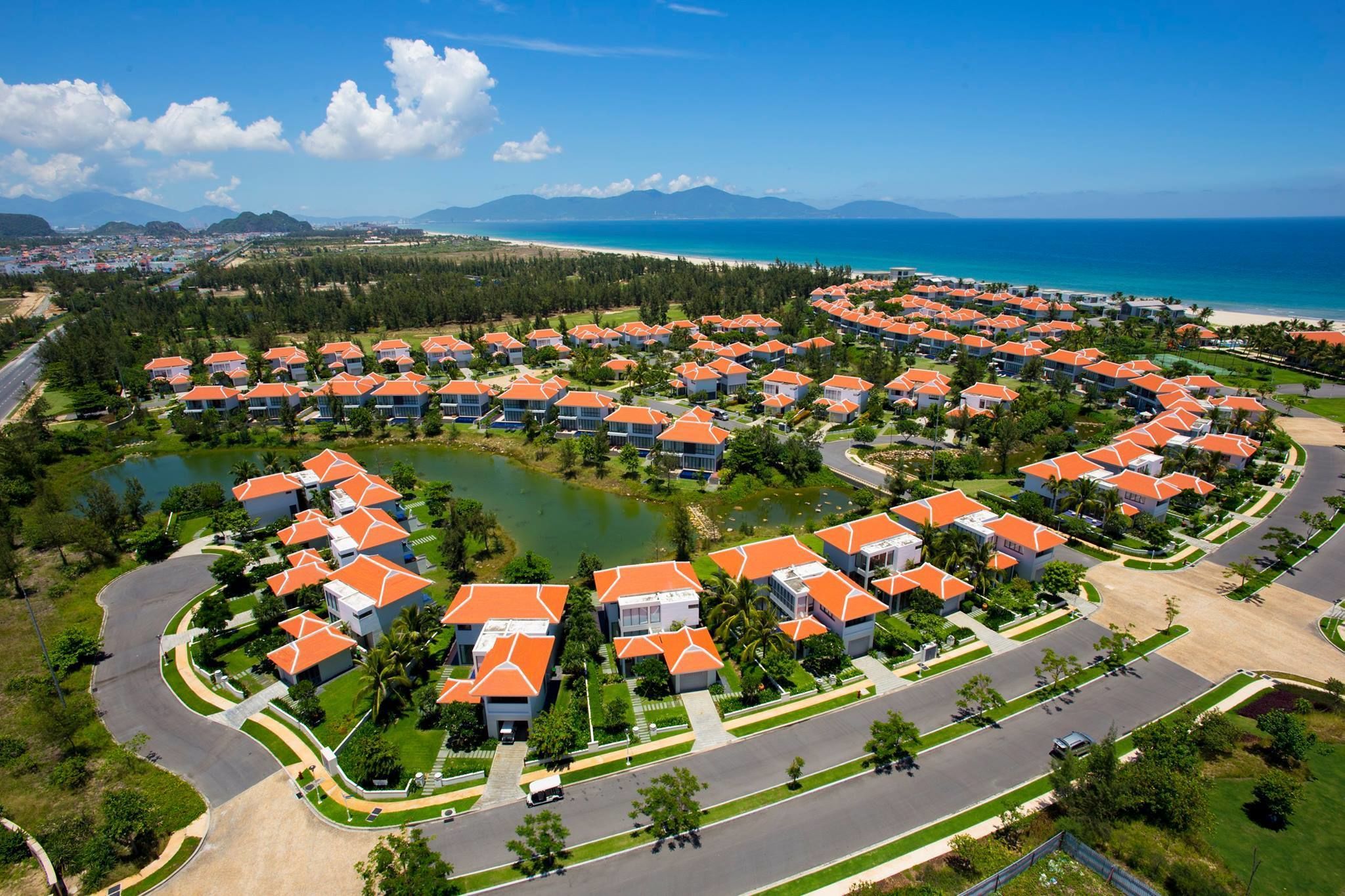 Vista aérea de un resort con varios edificios de techos naranjas, dispuestos en forma circular alrededor de una masa de agua central, con una playa y montañas al fondo.