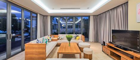 A modern living room with a sofa set, a coffee table, ottomans, a TV on a wooden stand, and large windows with curtains.
