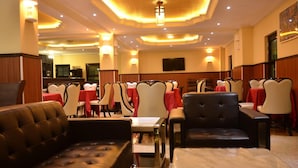 Un hall de hotel con una zona de estar, mesas con manteles rojos y un televisor en la pared.