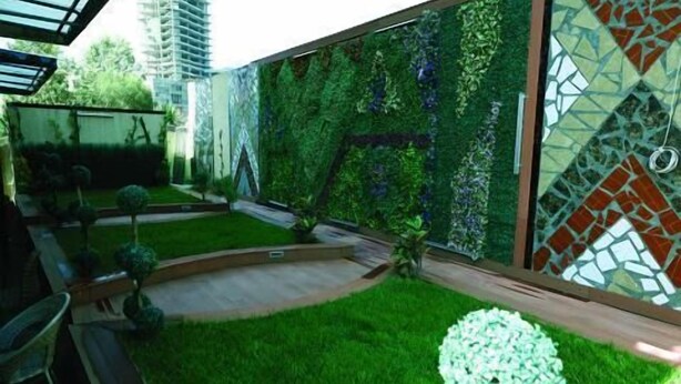Un espacio exterior moderno con un césped verde, un pequeño árbol y una pared decorativa con mosaicos.