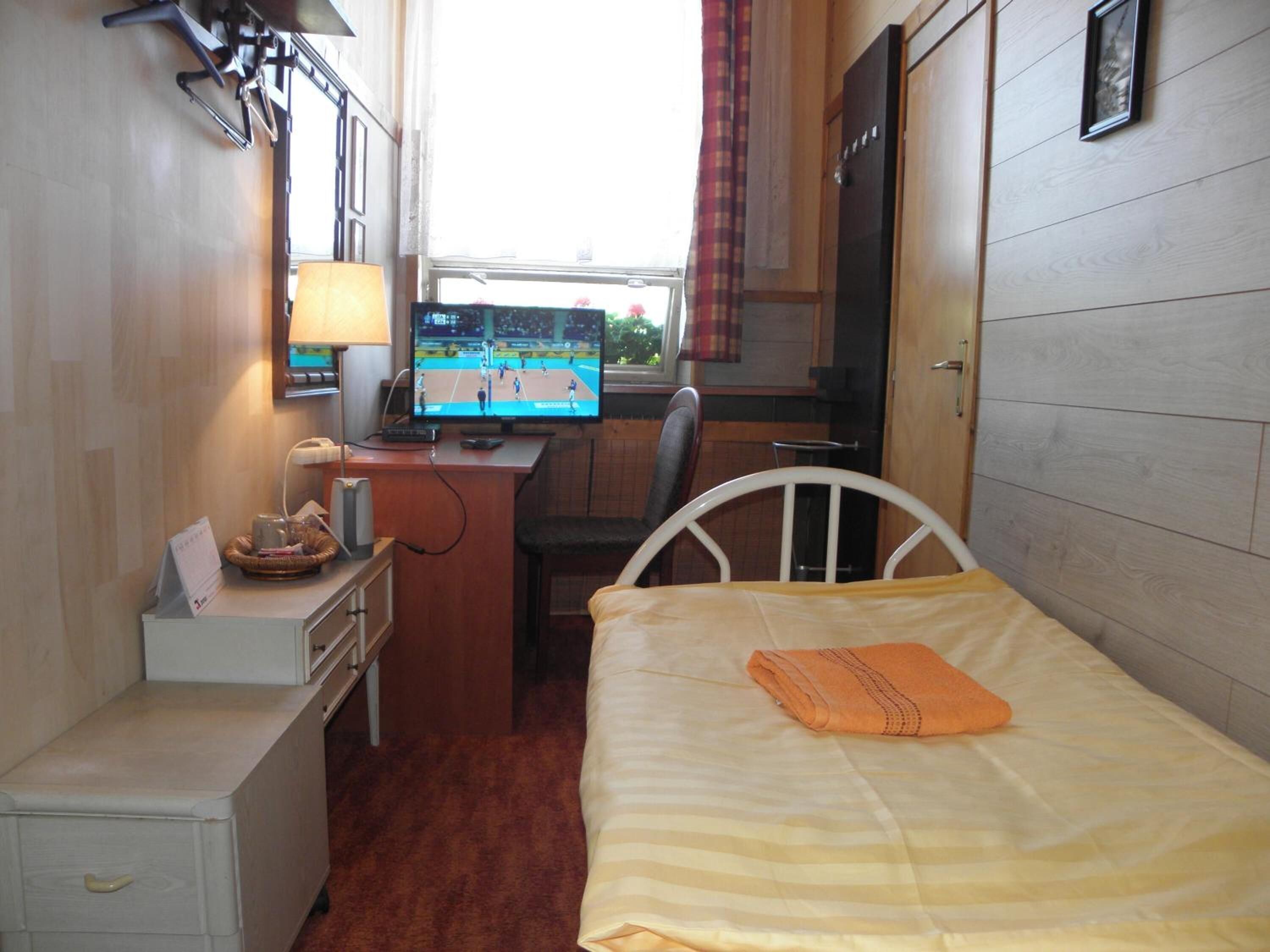 Una habitación de hotel pequeña con una cama, un escritorio con ordenador, una silla y un ventanal con cortinas.