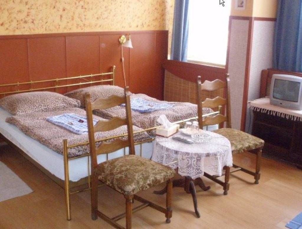 Una habitación con una cama, dos sillas de madera, una mesita redonda con mantel de encaje, un televisor y una ventana con cortinas.