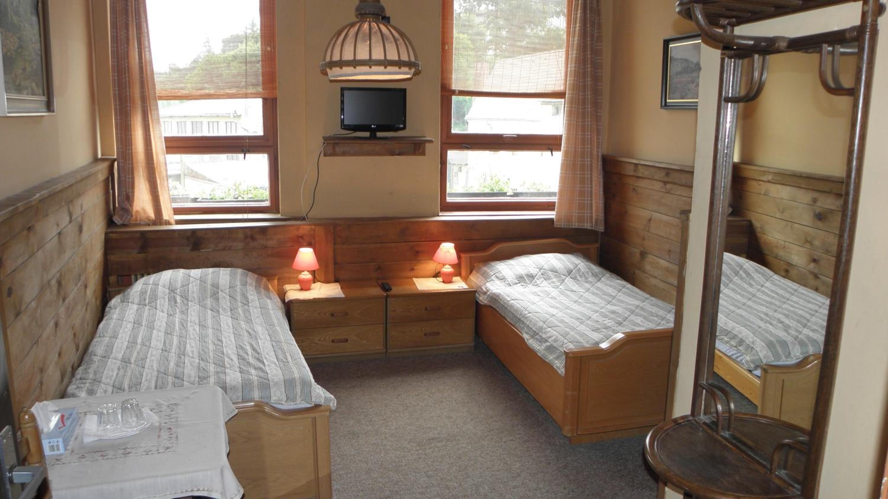 A room with two wooden beds, a small table, a mirror, and a television on a shelf.