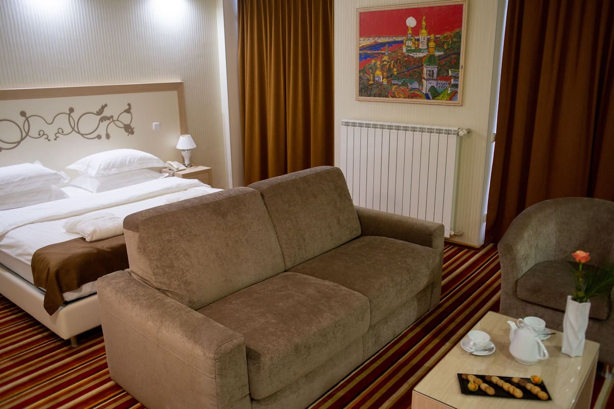 A hotel room with a bed, a sofa, a chair, a table with a teapot, cups, and a plate of pastries.