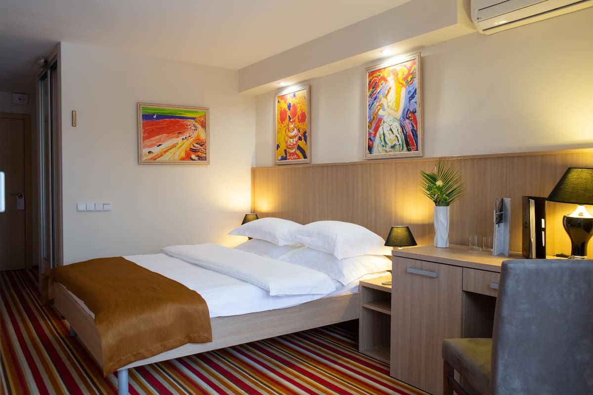 A hotel room with a bed, bedside tables, a desk, and a chair. There are paintings on the wall and a potted plant on the desk.
