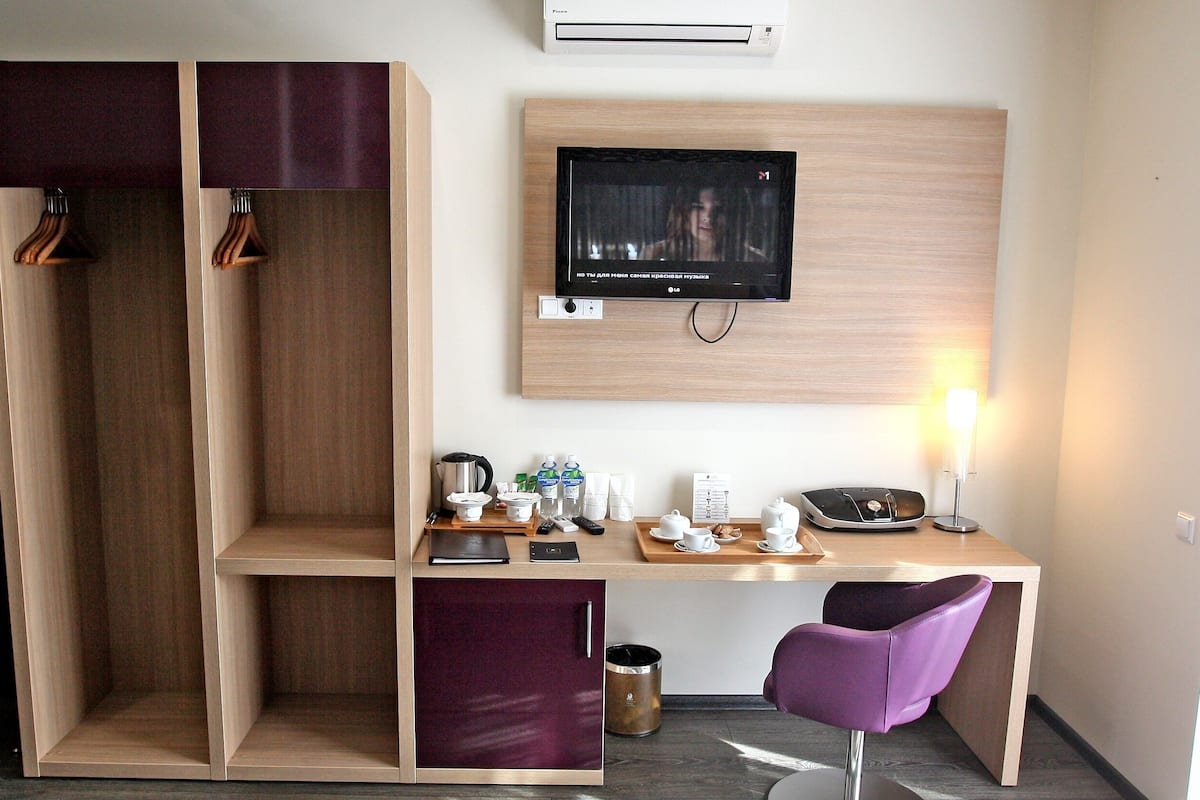 A compact hotel room with a wooden desk, a television, a purple chair, and a wardrobe with hanging space.