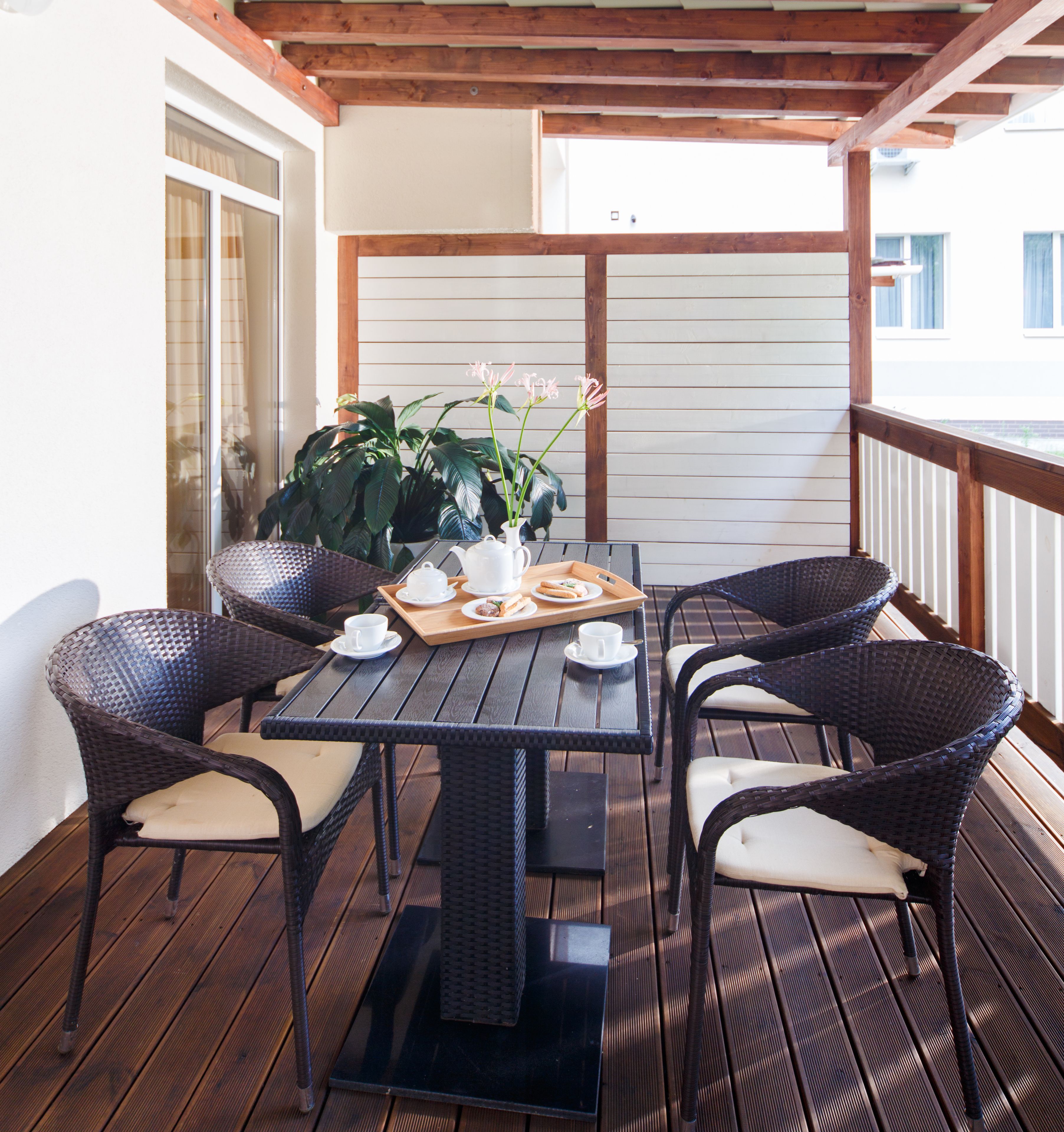 Una terraza de madera con una mesa, sillas, una planta en maceta y una bandeja con tazas y comida.