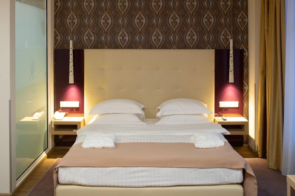 A hotel room with a large bed, two bedside lamps, a patterned wallpaper, and a glass shower enclosure.