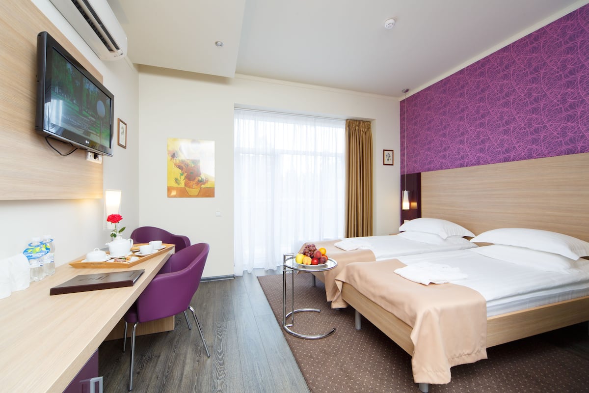 A hotel room with a bed, a desk with a chair, a small table with fruit, and a TV mounted on the wall.