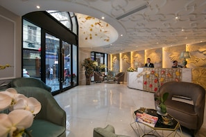A modern hotel lobby with a reception desk, seating area, and large windows offering views of the street.