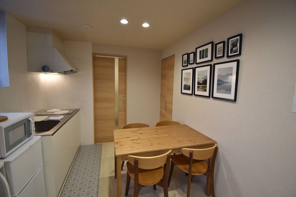 A compact kitchen with a microwave, sink, and wooden cabinets. There is a dining table with four chairs and a small rug.