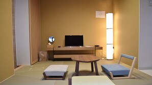 A hotel room with tatami flooring, a low wooden desk with a flat-screen TV, a small round table, two tatami mats, a blue chair, and a wall-mounted lamp. - ieLIFE Resort HIGASHIYAMA (Kyoto)