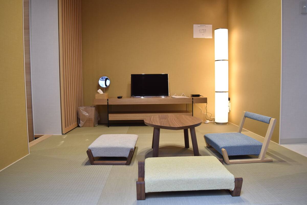 A hotel room with tatami flooring, a low wooden desk with a flat-screen TV, a small round table, two tatami mats, a blue chair, and a wall-mounted lamp.