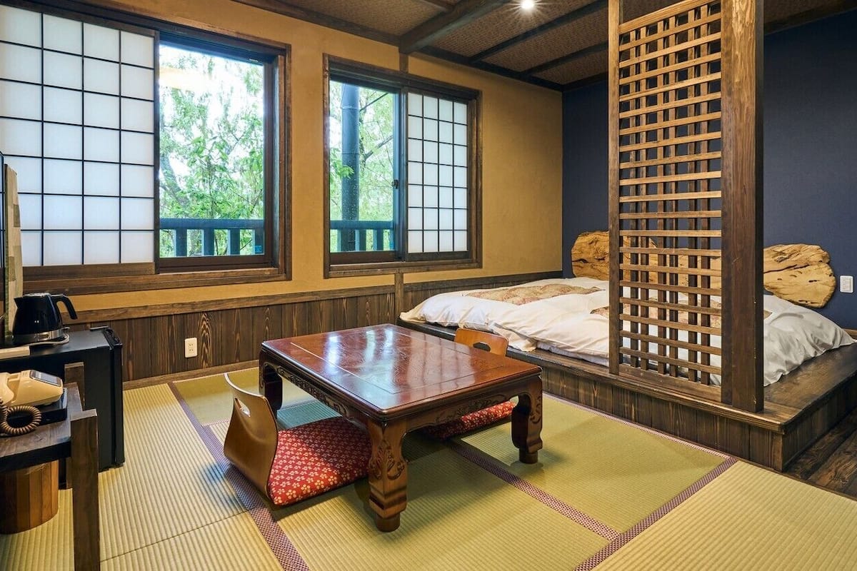 A traditional Japanese room with tatami flooring, a low wooden table, a bed with a wooden headboard, and sliding doors with a view of greenery outside.