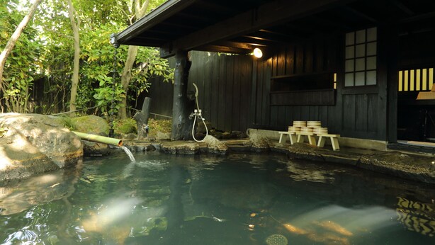 Un onsen japonés tradicional con una estructura de madera, una fuente termal y un camino de piedra.